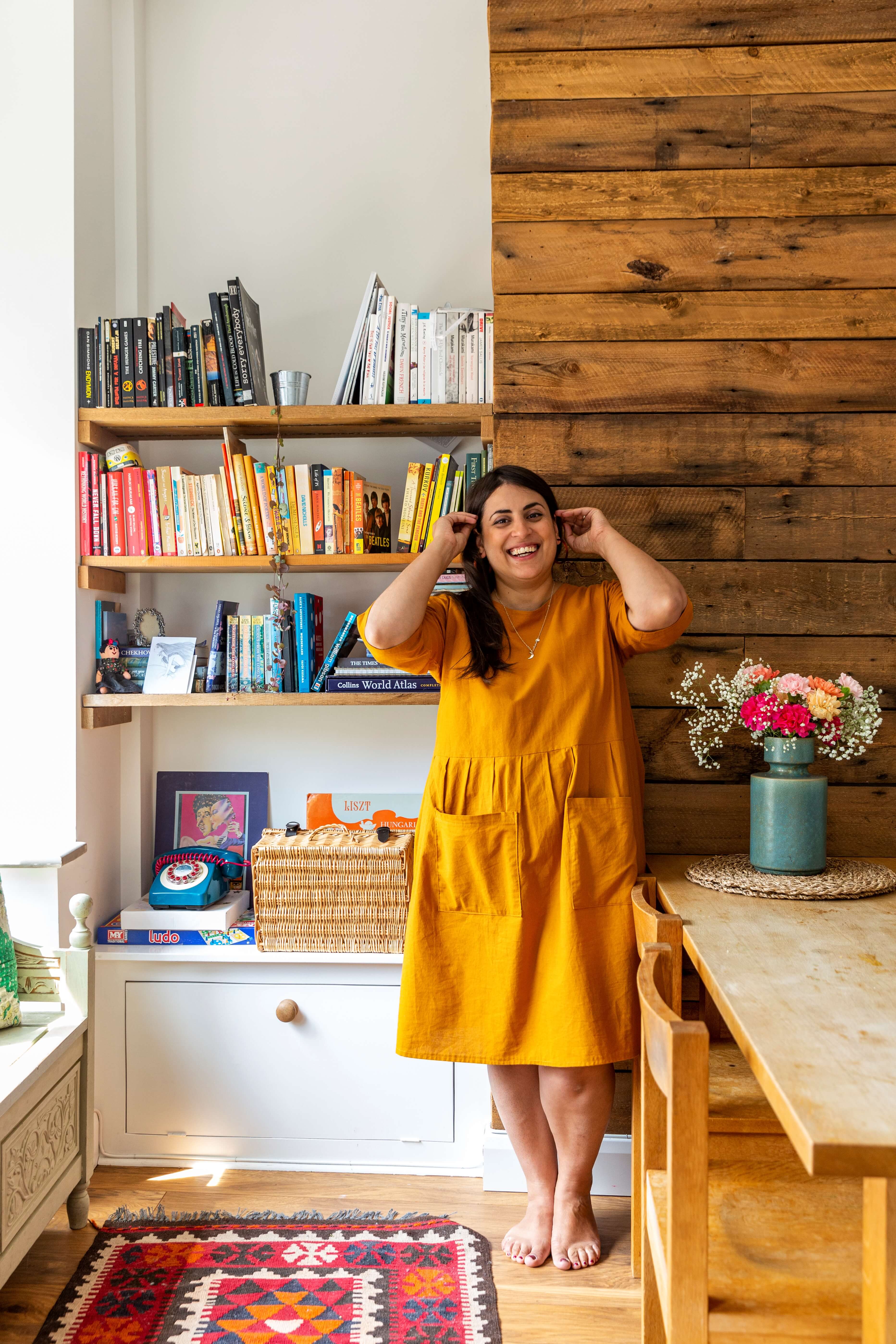 Rida is standing in front of the bookshelf wearing a mustard yellow dress and laughing