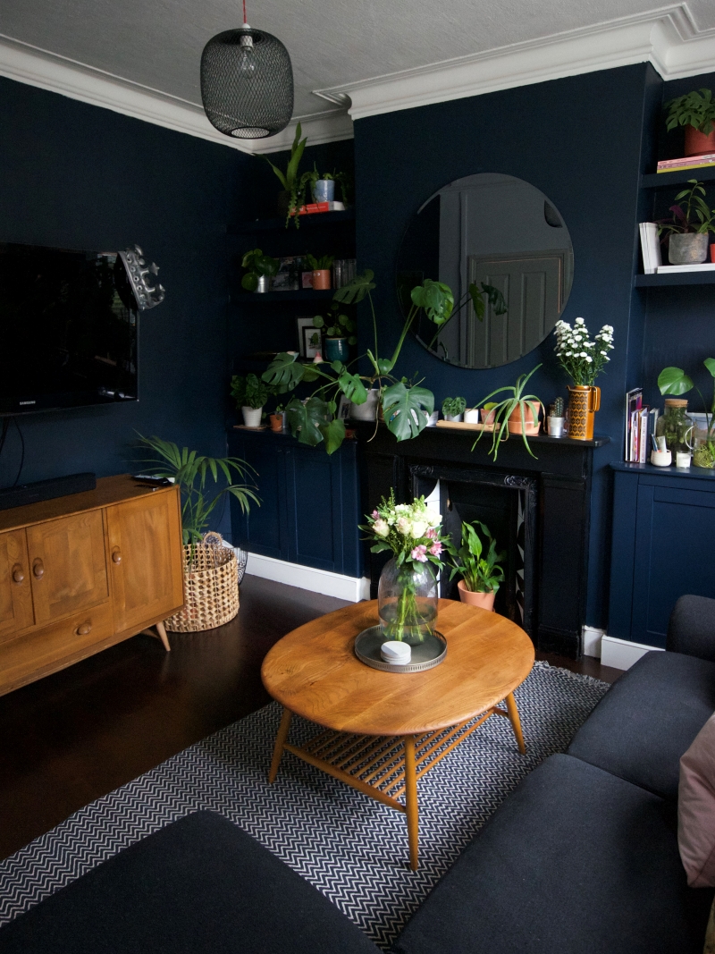 Dark painted wall with dark carpets decorated with floral cushions, mid century furniture and plants 