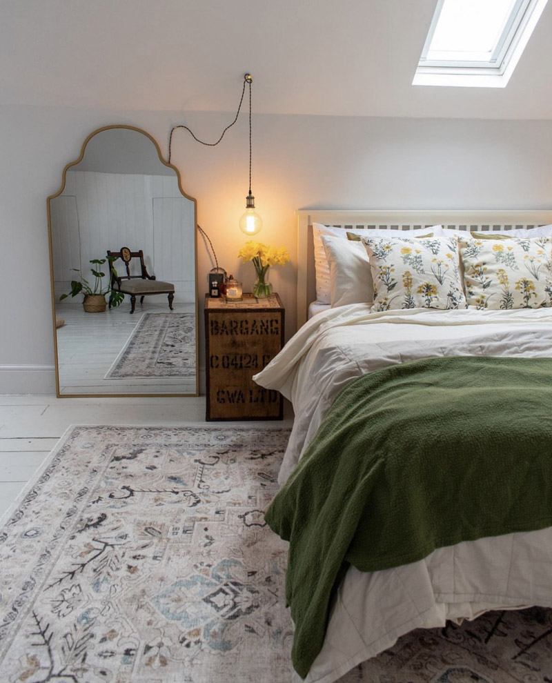 A white fresh loft room, with green and subtle floral bedding, vintage arched mirror leans on the wall and next to a crave bedside table a low vintage bulb hangs low next to the bed.