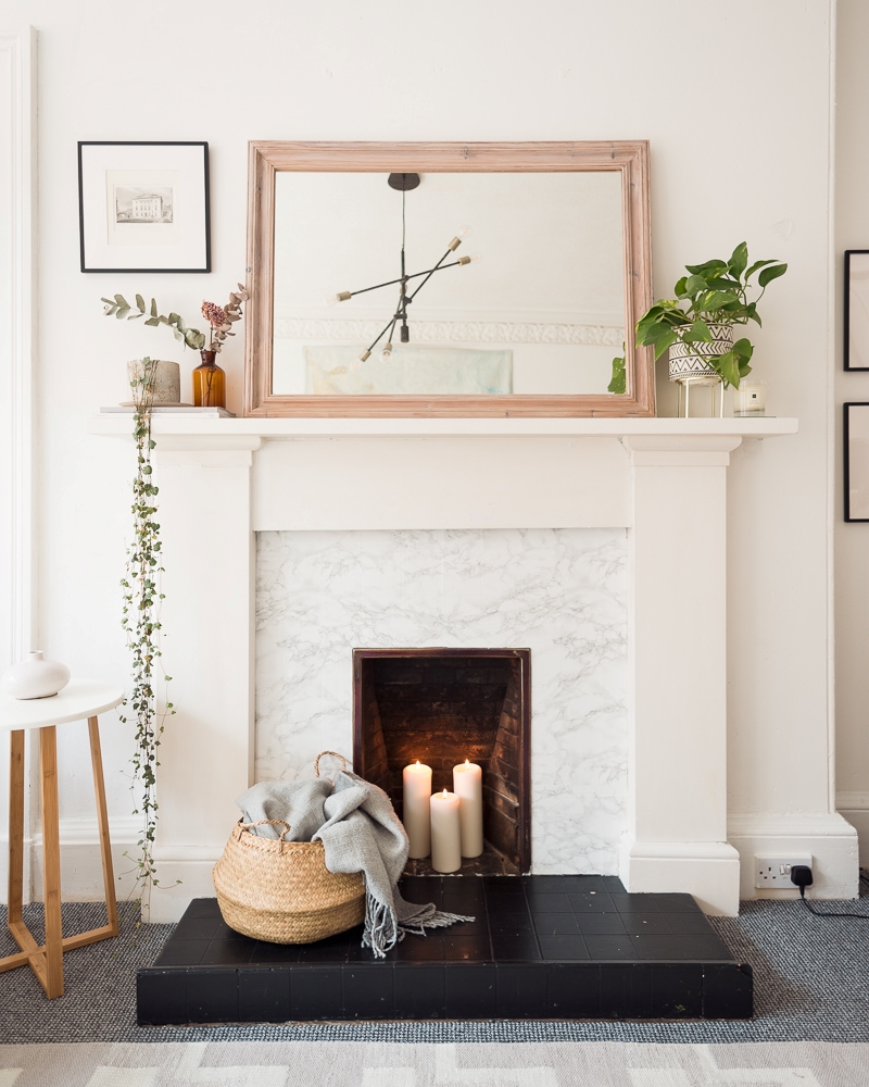 Cosy candle lighting in a faux fireplace, a belly basket with a grey blanket is in, plants sit on the mantle and a large neutral mirror. 